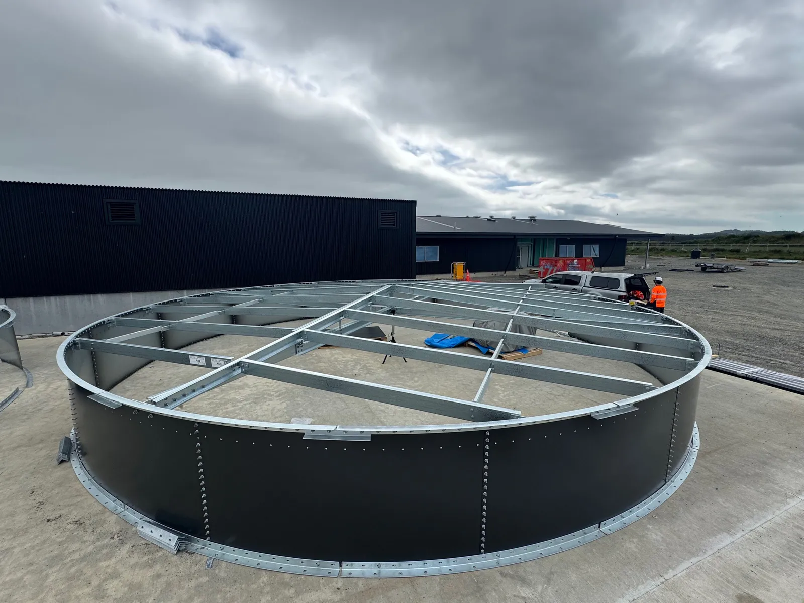 The roof steel frame bolted together at ground level inside the first ring of a tank shell, on the concrete pad beside the site buildings