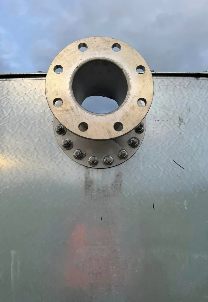 A bolted outlet flange through the top of a galvanised tank wall, seen against an overcast sky