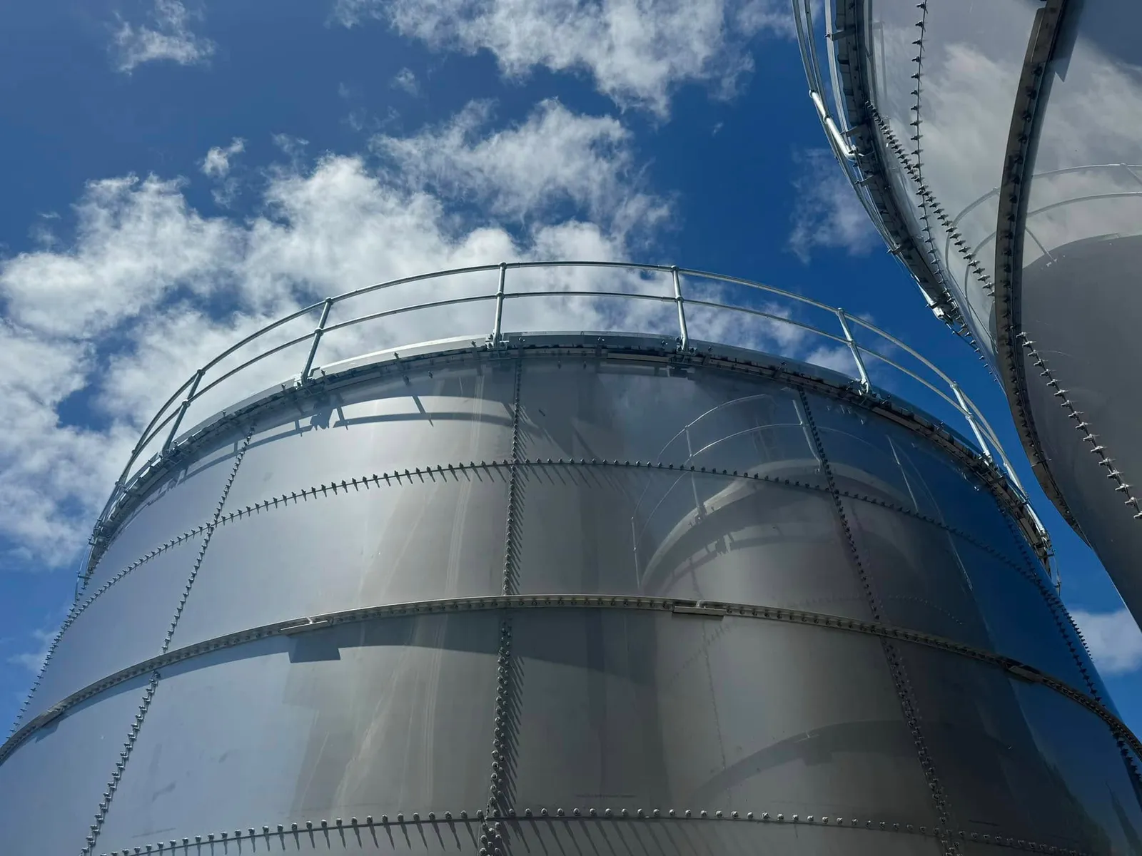 Looking up the wall of a galvanised bolted steel tank to the handrail around its rim, a second tank alongside