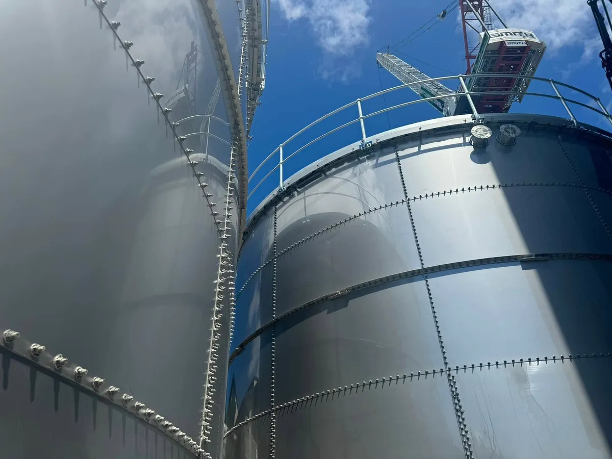 Looking along the wall of a tall galvanised bolted tank, with the roof handrail running away against a blue sky