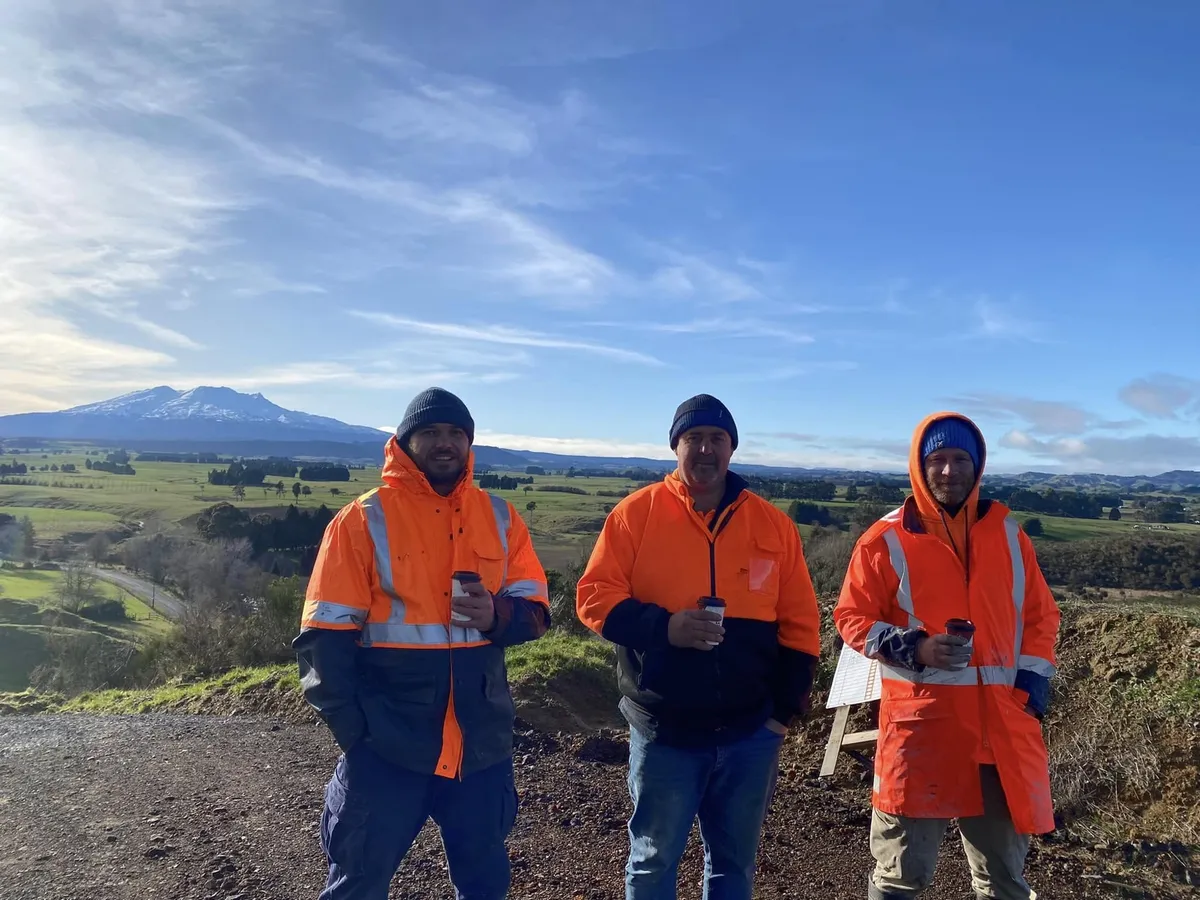 Three Impact crew in hi-vis jackets on a gravel access road, with farmland and a snow-capped range behind them