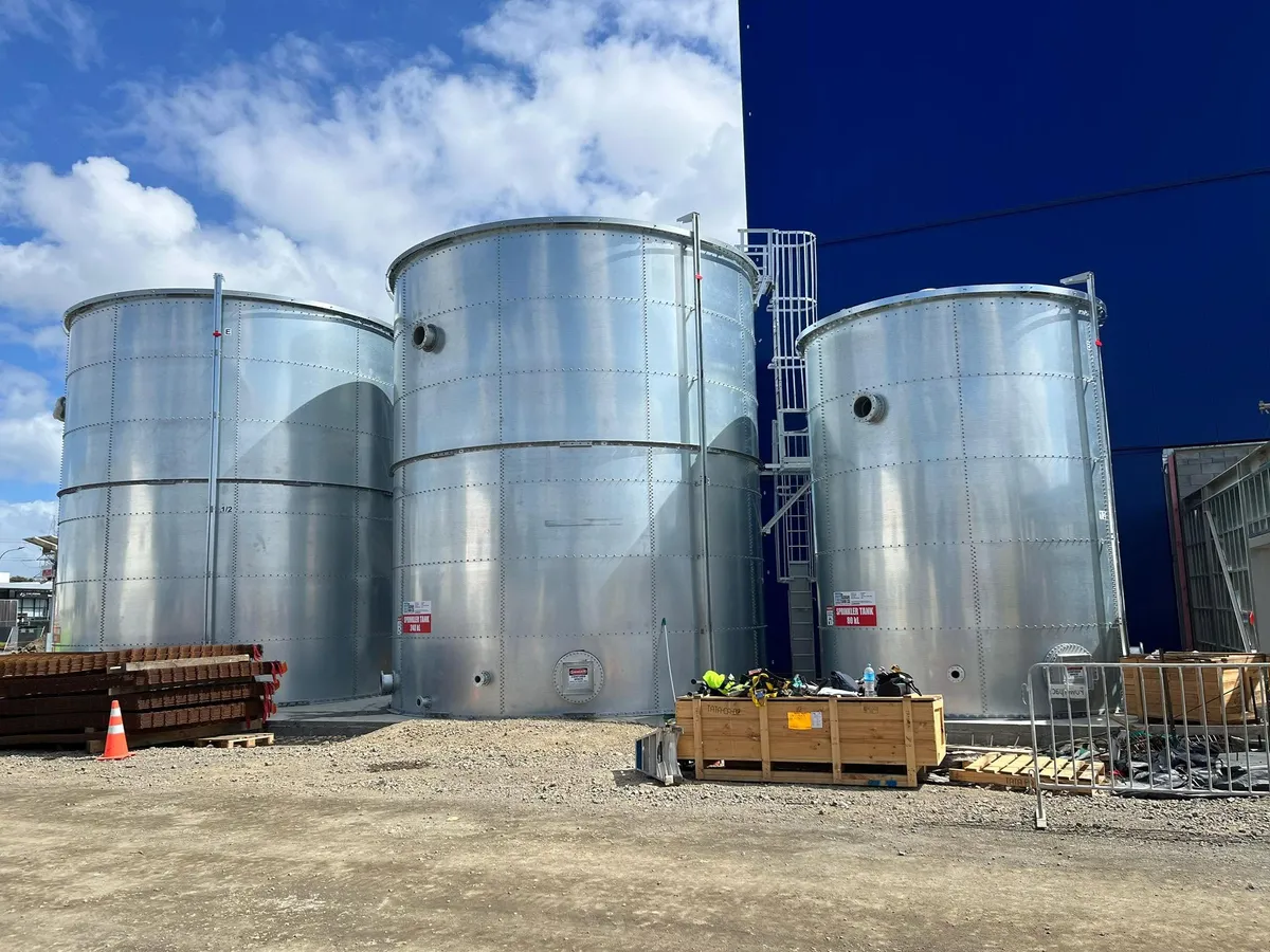 Three galvanised bolted tanks of differing heights standing together on a site under cloud