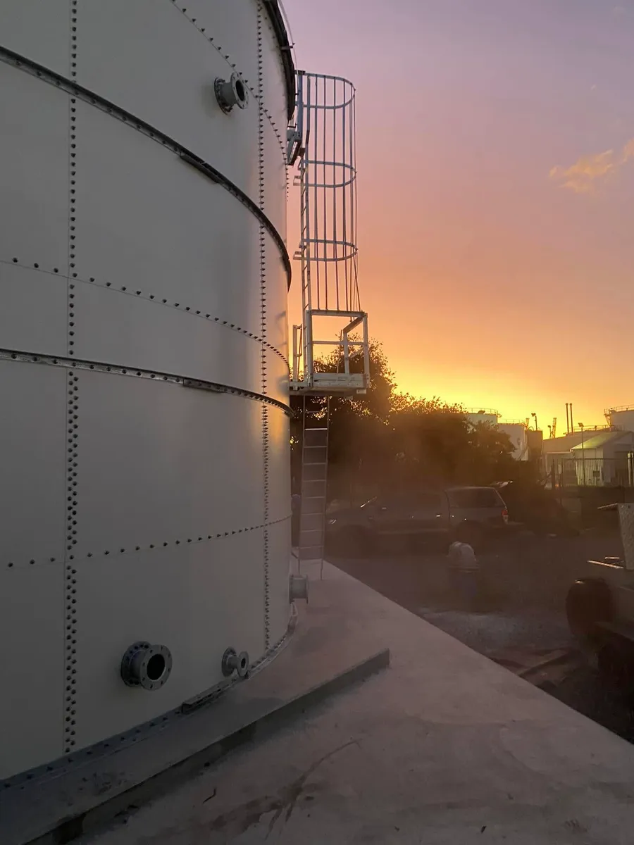 A white bolted steel tank and its caged access ladder against an orange sunset sky