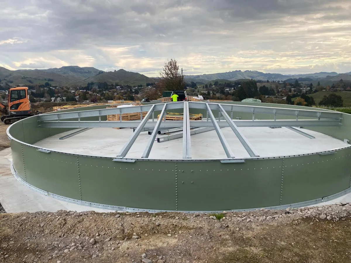 Radial roof steelwork spanning the open top of a green tank shell, hills behind